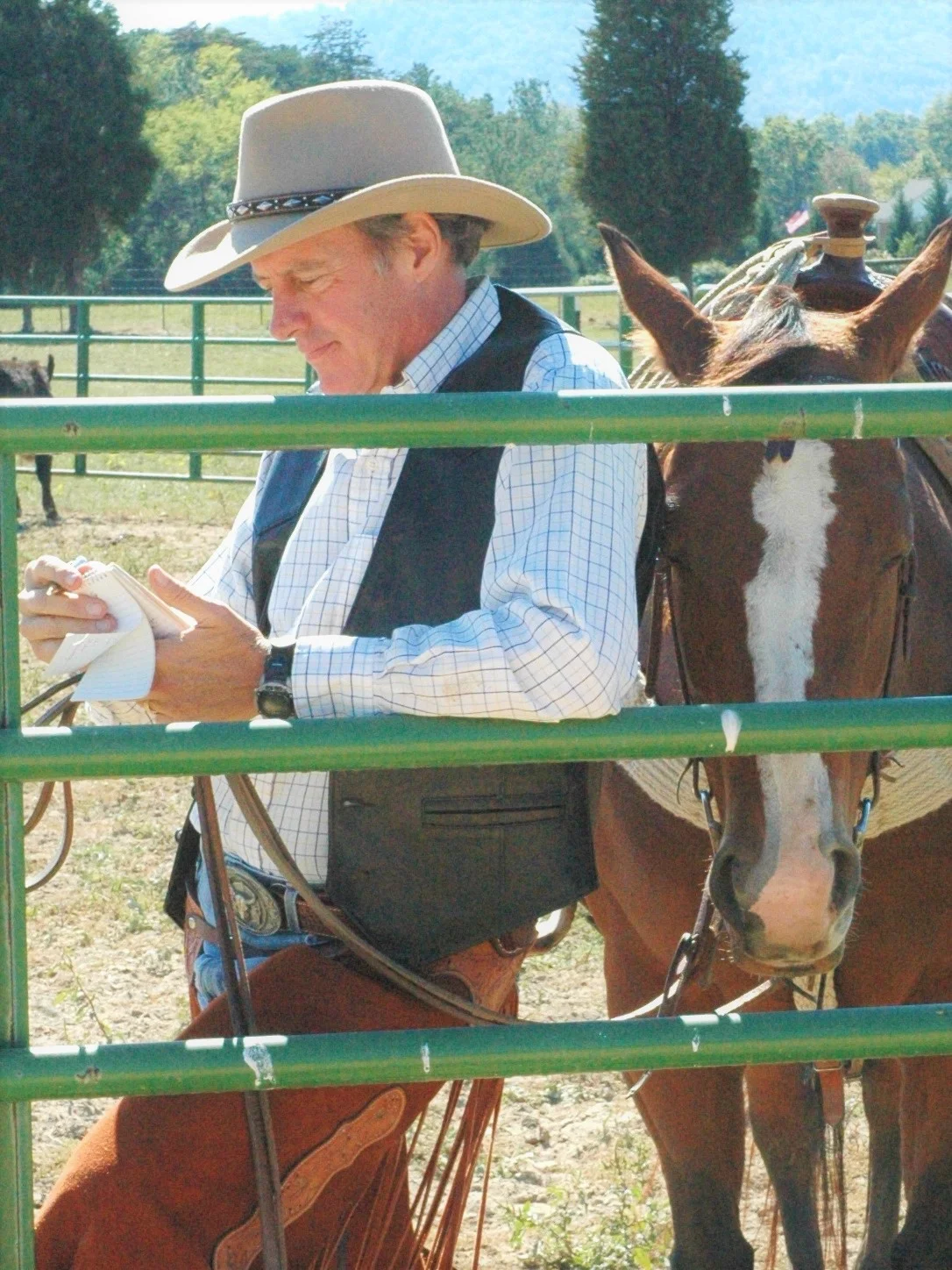Michael J. Hightower in western attire — cowboy hat, vest, and chaps — standing beside a brown-and-white horse at a green ranch fence on a sunny day