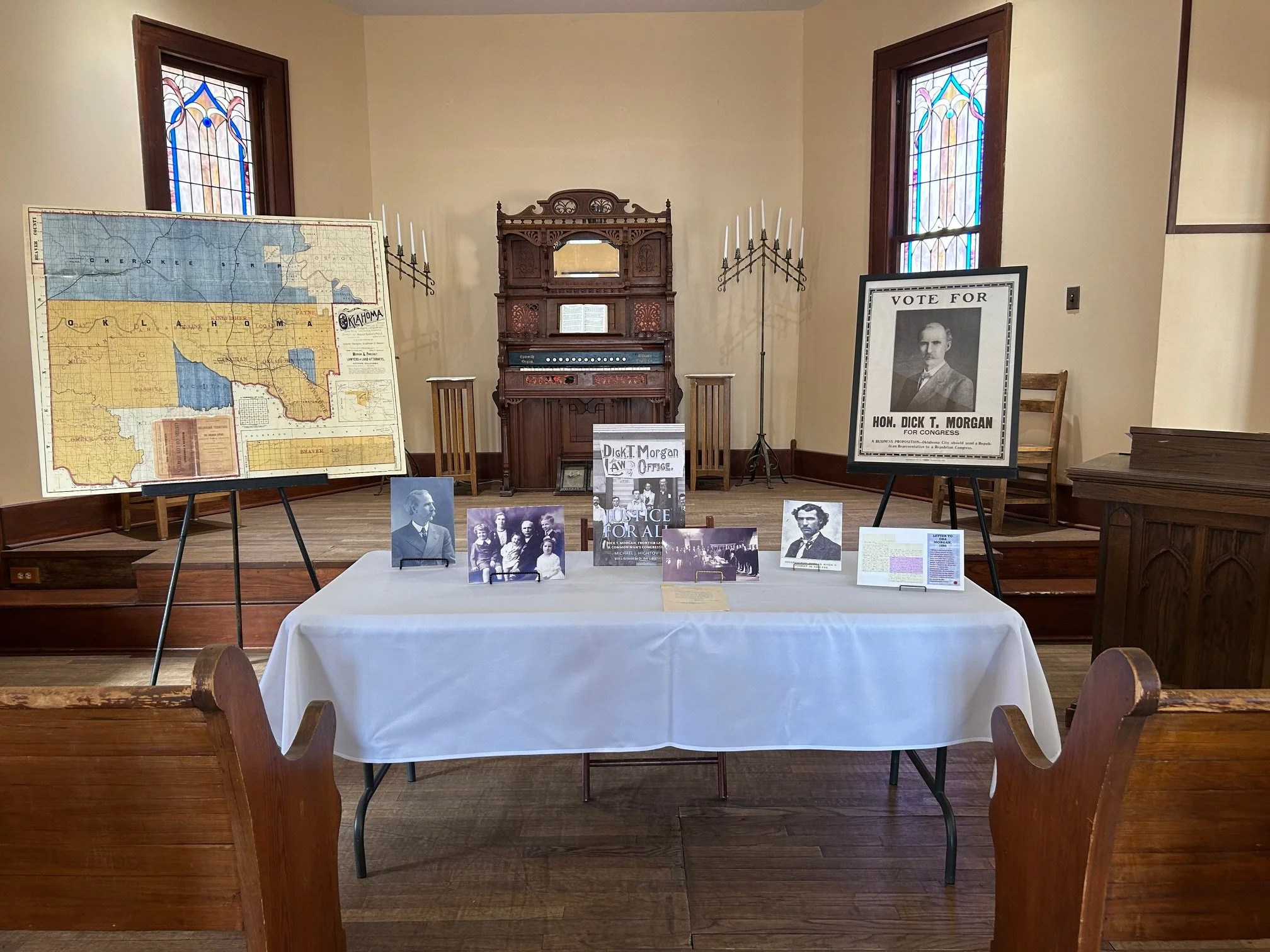 Display table set up in a historic chapel room at a February 22, 2025 event, featuring books, historical photographs, a vintage Oklahoma map, and a Vote for Hon. Dick T. Morgan campaign poster on an easel