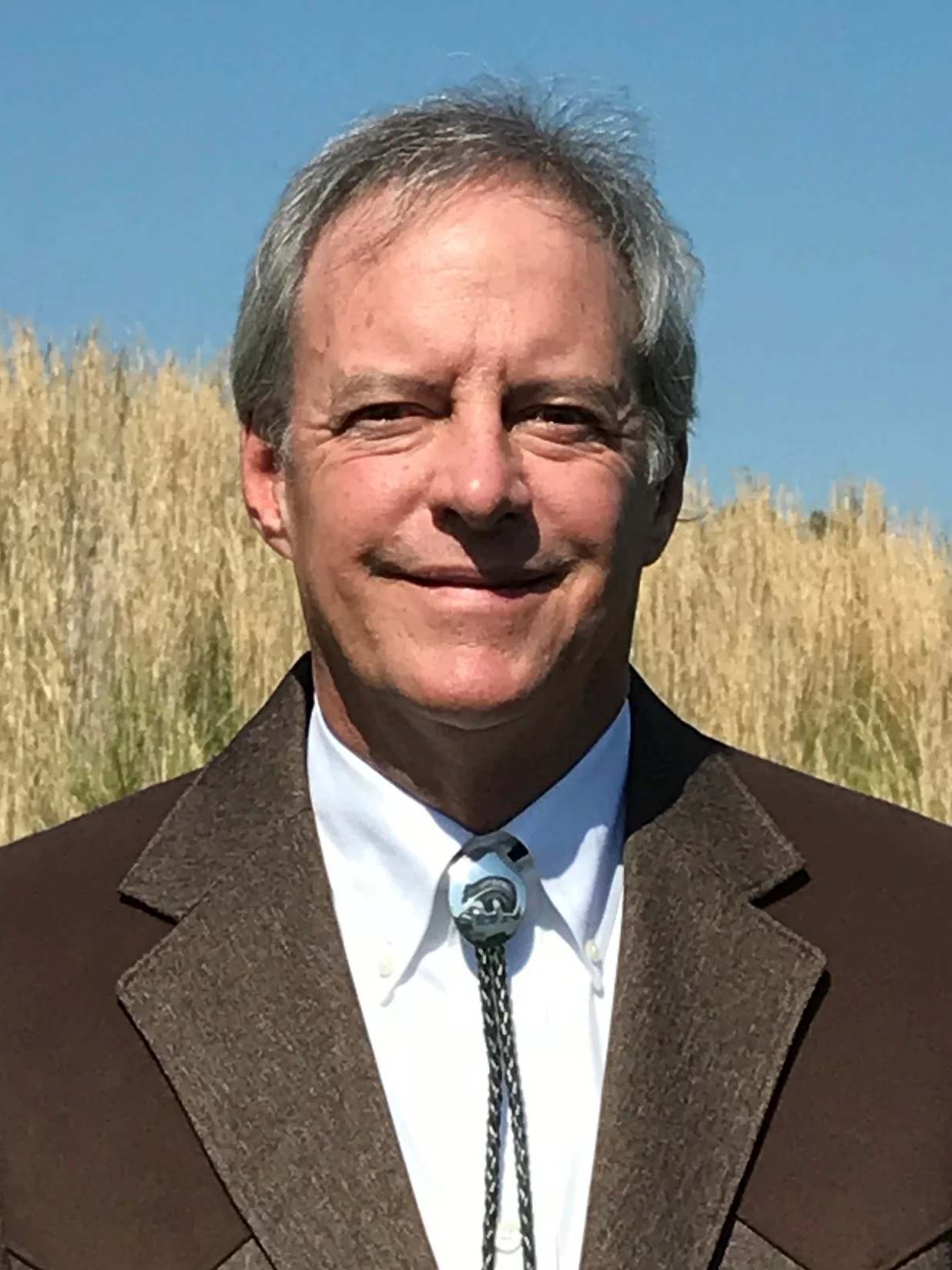Michael Hightower wearing a brown blazer, white shirt, and bolo tie, smiling in front of tall golden grass under a blue sky.