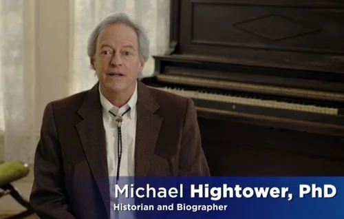 Video still of Michael Hightower, PhD, identified on screen as Historian and Biographer, speaking in front of a vintage upright piano